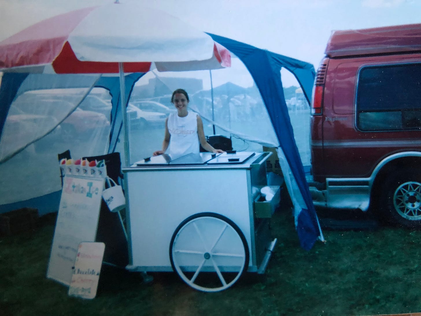 A young person smiles and stands behind a mobile gelato cart, sheltered by a red and white umbrella