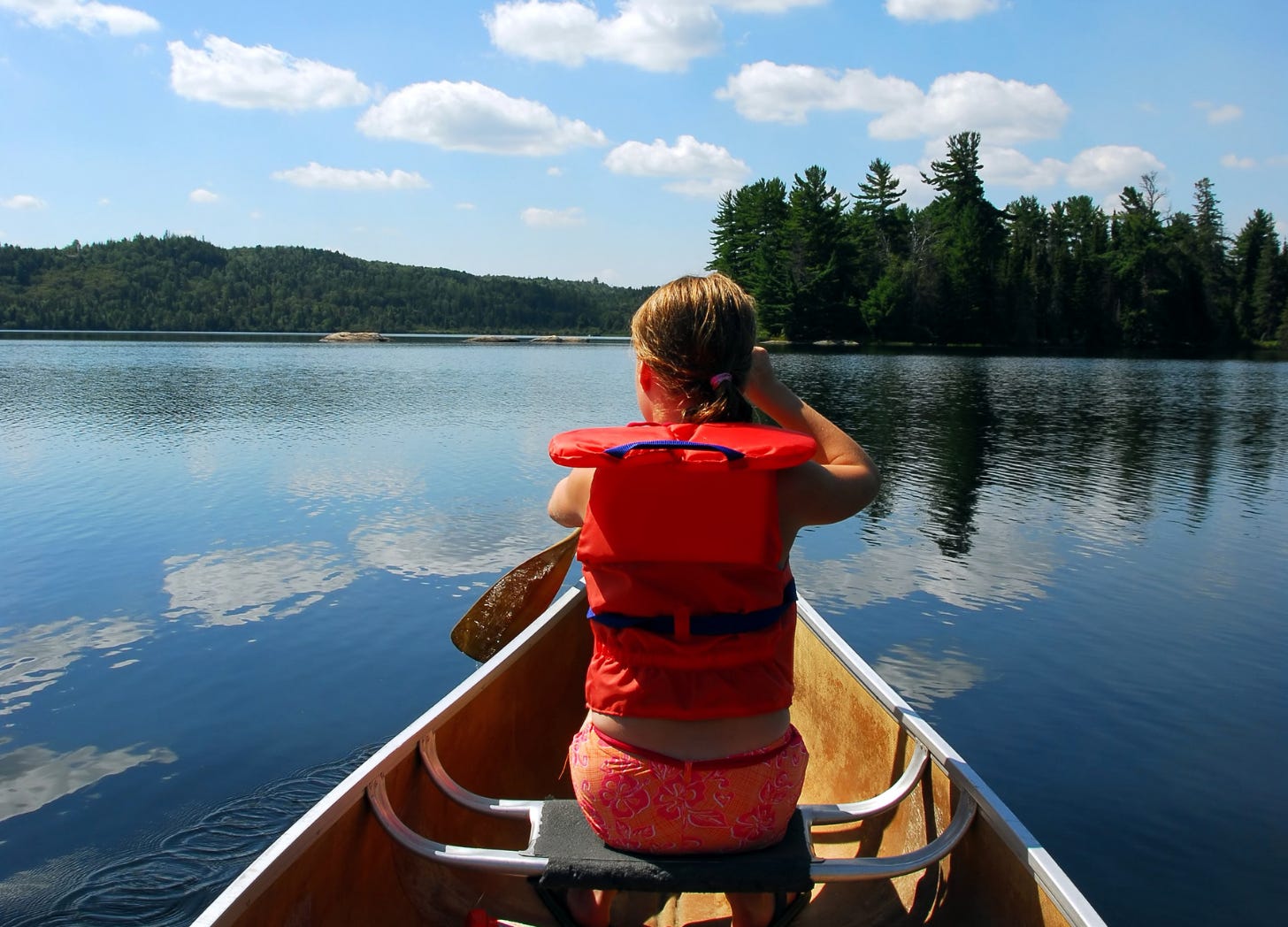 young girl paddles a canoe alone on calm water, wearing a lifejacket. The view is from behind as she heads toward a small island in the distance. young girl paddles a canoe alone on calm water, wearing a lifejacket. The view is from behind as she heads toward a small island in the distance.
