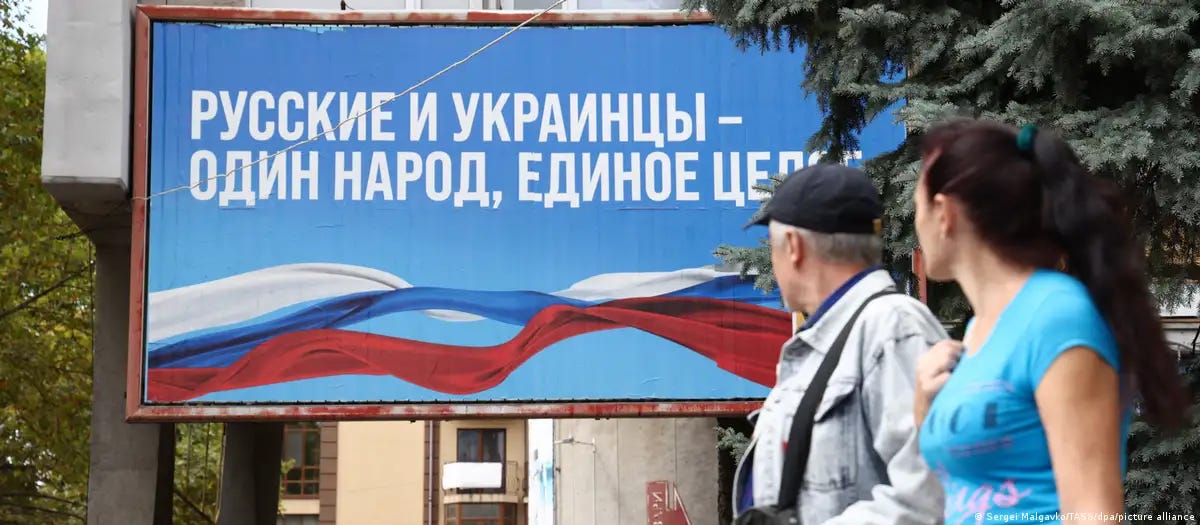 A man and woman looking at a sign that reads, “Russians and Ukrainians are one people, one whole.”