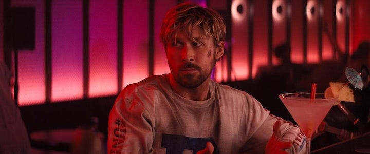 Ryan Gosling sitting in a neon-lit bar, wearing a worn long-sleeve shirt and looking confused while holding a comically large cocktail with an umbrella and garnish.