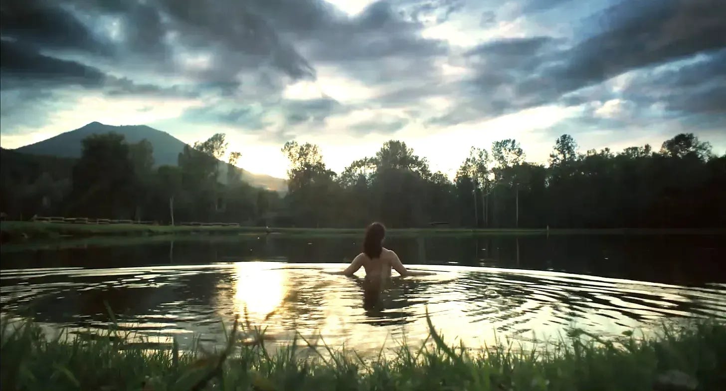 A person wades nude into a still lake at sunset, surrounded by grass, trees, and distant mountains. The sky is moody with dark clouds streaked by golden light, and the water reflects the dramatic colors of the evening.