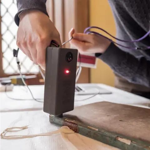 University of St Andrews A man's hand holding a plastic gun-like object against a green covered book. The man is wearing a blue jumper.