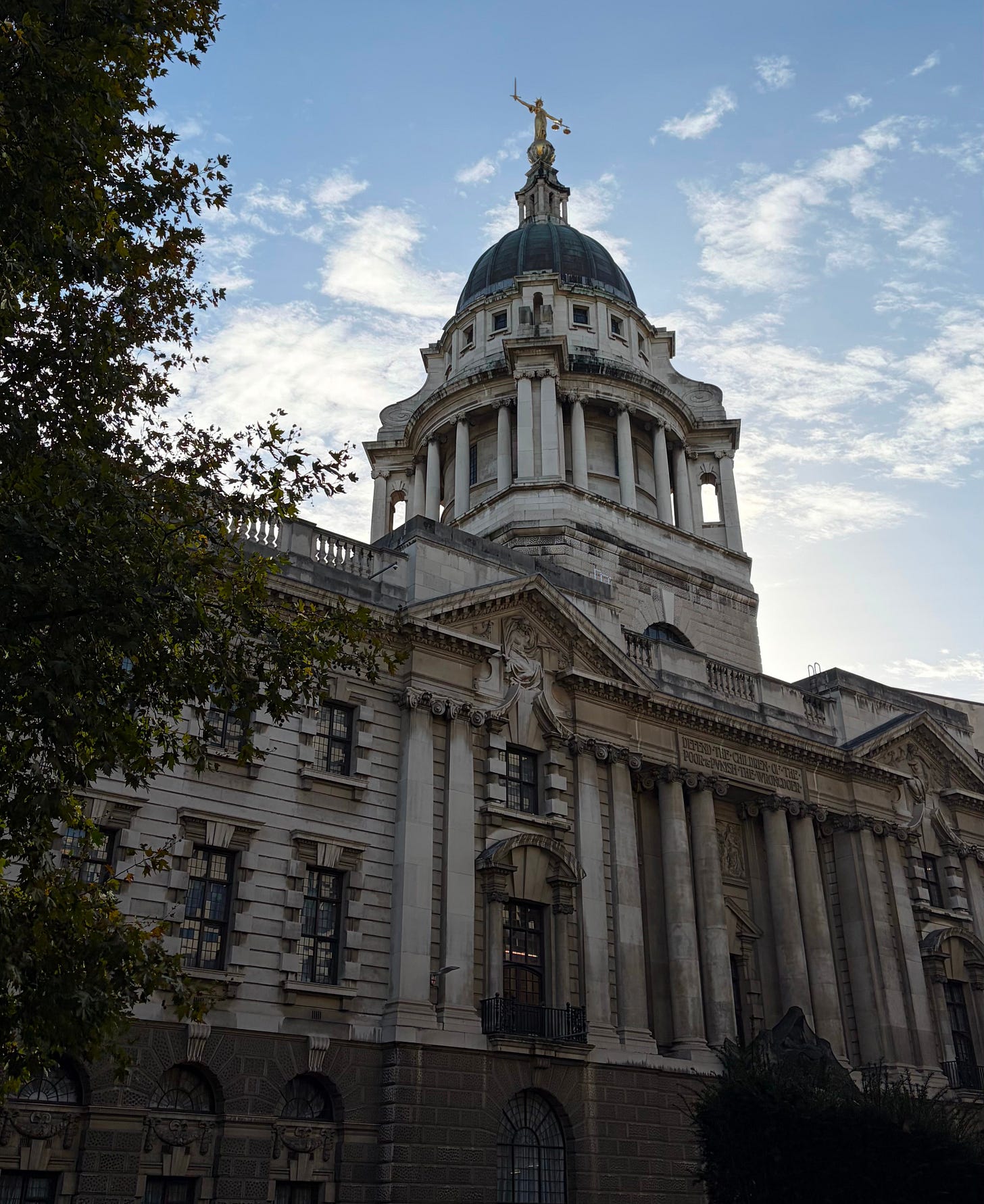 A large historic building with classical architecture, featuring tall columns and ornate stone detailing. A domed roof rises at the centre, topped with a gold statue holding scales and a sword. The building is photographed from a low angle against a blue sky with scattered clouds, with tree branches framing the left side. A large historic building with classical architecture, featuring tall columns and ornate stone detailing. A domed roof rises at the centre, topped with a gold statue holding scales and a sword. The building is photographed from a low angle against a blue sky with scattered clouds, with tree branches framing the left side.