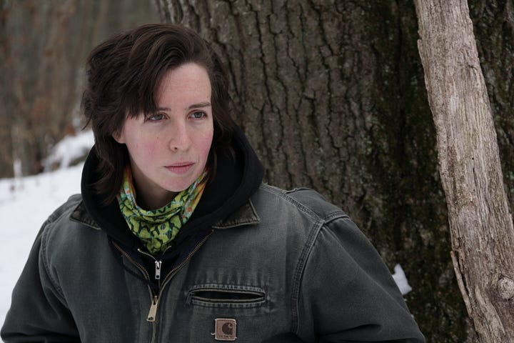 the author, a masculine white person with short brunette hair, stands outside by a tree; snow blankets the background. next, the camera peers over their shoulder as they write in a notebook at a desk that oversees the wintery landscape