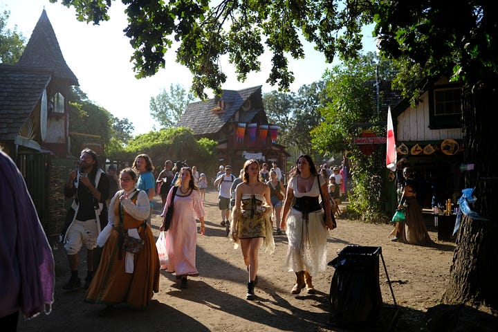 Bards singing in a tavern wearing medieval garb, an elf and wizard surveying goods at a shop, fire-twirler show, Geralt and another warrior deep in conversation, a man playing dead with an arrow under his armpit, guests walking toward the camera, a woman with a horse head looking over a table.