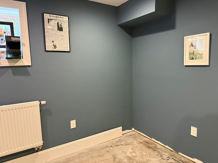 Alt text: A four-photo collage documenting the construction of a custom built-in reading nook. The top-left image shows an empty corner of a room with blue walls, white trim, and a ceiling bulkhead. The top-right image shows the initial framework for a bench seat and a vertical cabinet side installed beneath the bulkhead. The bottom-left image shows the bench structure further along, with additional framing and support panels in place. The bottom-right image shows the nearly completed project, including a built-in bench with storage compartments below and a floor-to-ceiling bookshelf fitted beneath the bulkhead. Together, the images illustrate the progression from an empty corner to a custom built-in seating and storage feature.