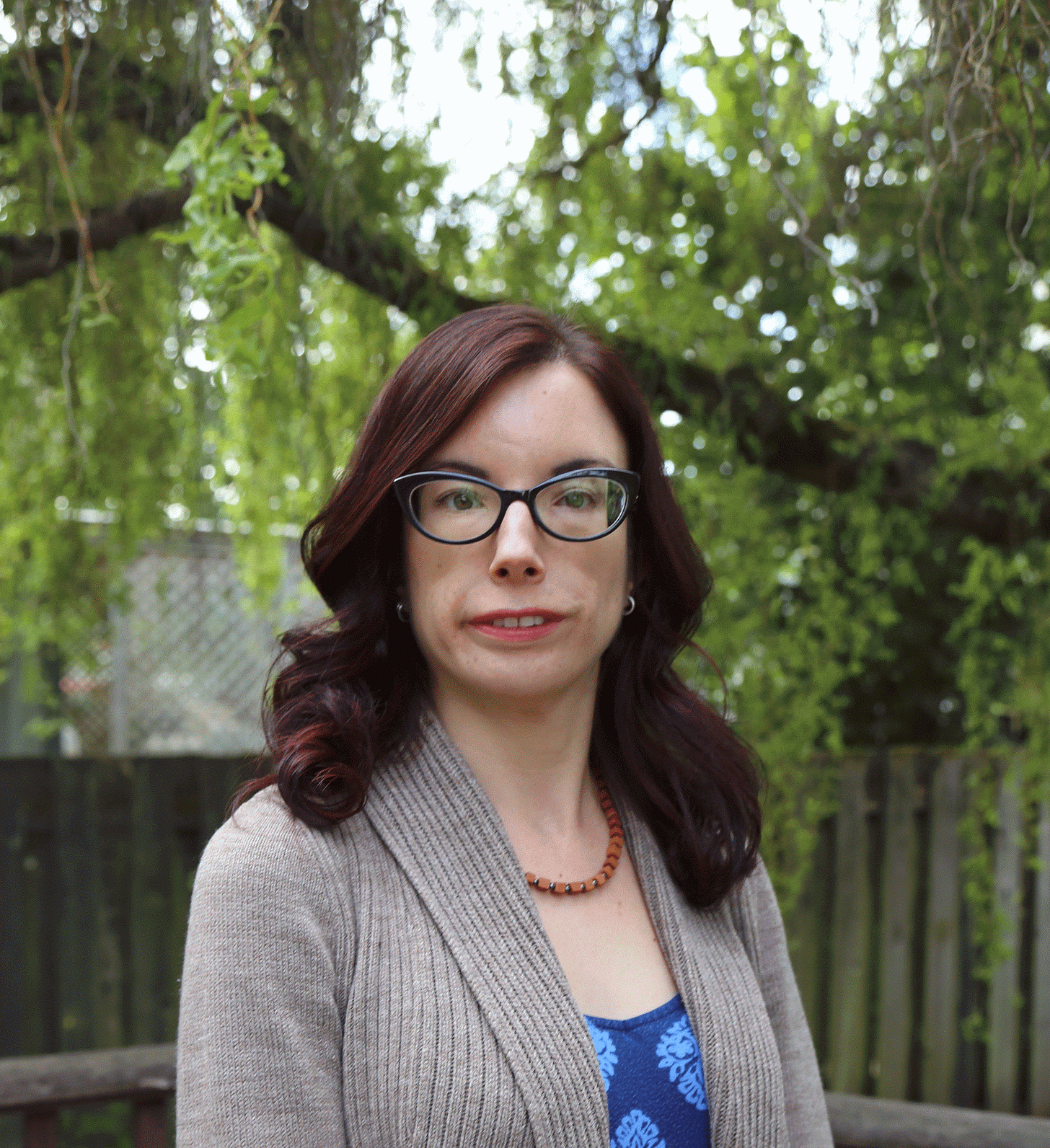 A photo of a smiling white woman with long dark-auburn hair. She is wearing glasses, a blue shirt, a beige cardigan, and a necklace.