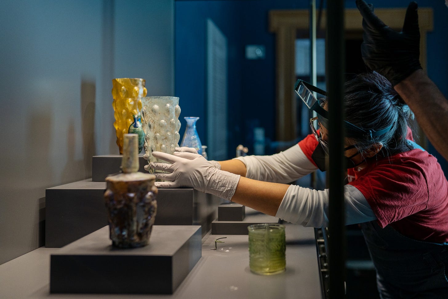 A person uses wire and wax to secure ancient glass objects in a museum exhibition case A person uses wire and wax to secure ancient glass objects in a museum exhibition case