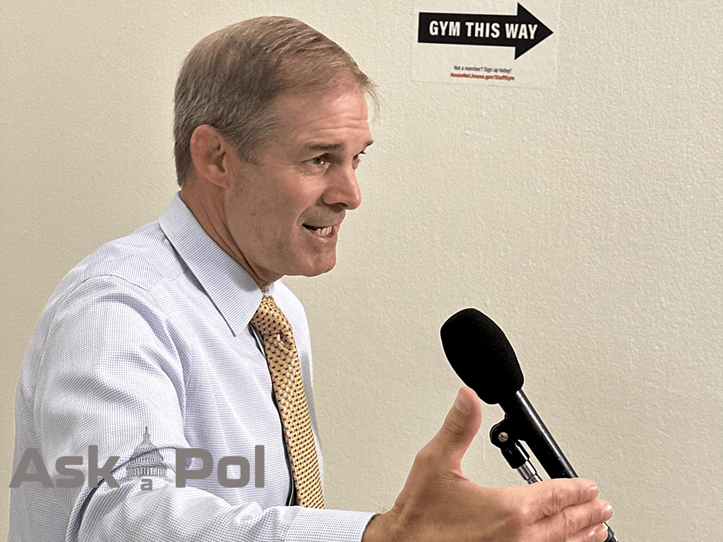 A man in a collared shirt and tie speaks into a microphone while waving his right hand. Photo: Matt Laslo for © www.askapolpolitics.com