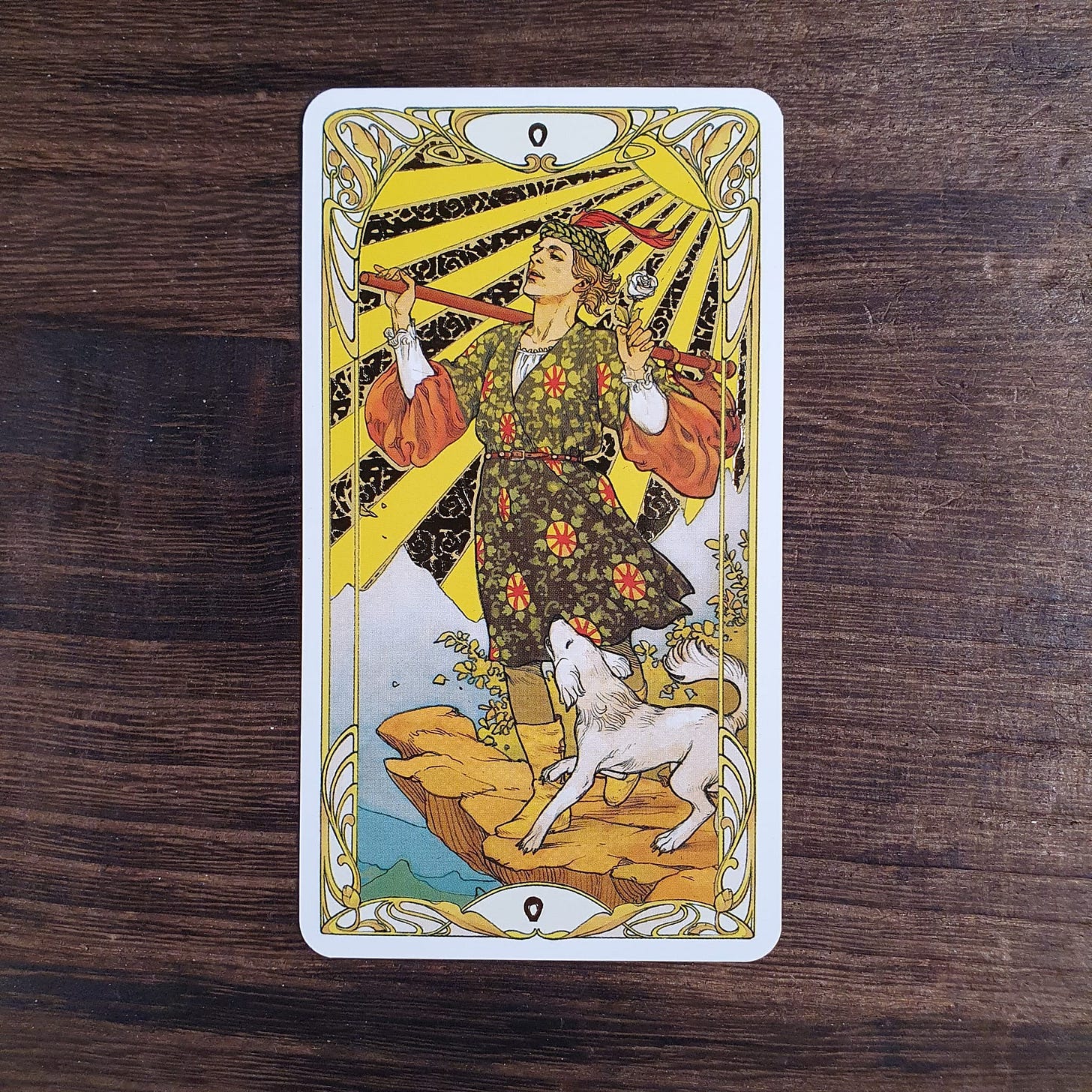 The Fool card from the Golden Art Nouveau Tarot held beside a small dog wearing a collar bell, photographed for the Tarot DMs interview with Alice Slater by Finbarre Snarey.