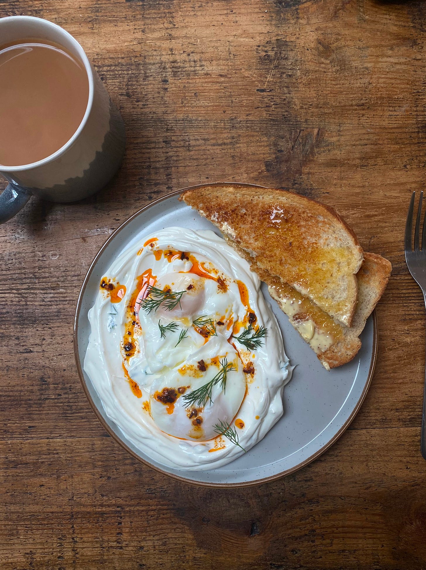 a plate on a wooden table. Next to the plate is a mug of tea. On the plate, there is thick yoghurt with poached eggs, dill, chilli oil and toast