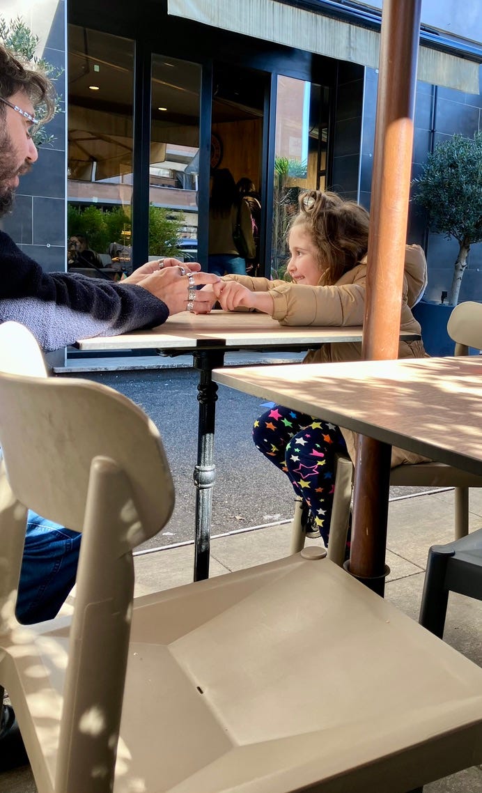 Papà e figlia seduti a un tavolino all'aperto si tengono le mani sorridendo, in una giornata di sole.