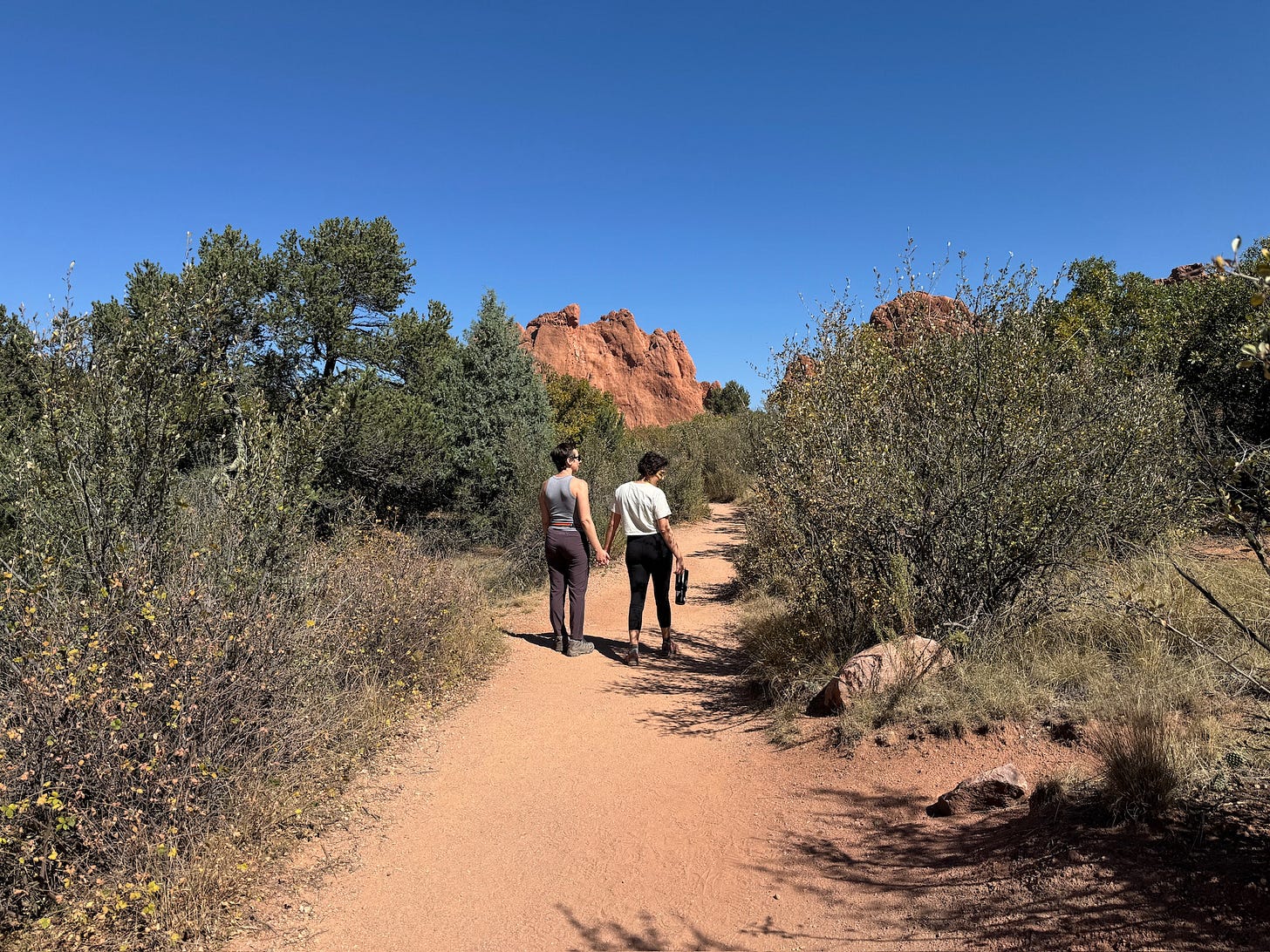 Sam and Erin hold hands while walking down a red dirt path, surrounded by desert shrubs, with a red rock formation in the background, and a clear blue sky overhead.