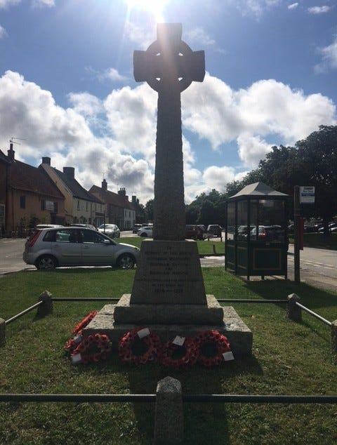 Village war memorial, stone cross with the names of the dead surrounded by a low fence, poppy wreaths laid against the stone plinth, in background there are cars, cottages, trees and a bus stop