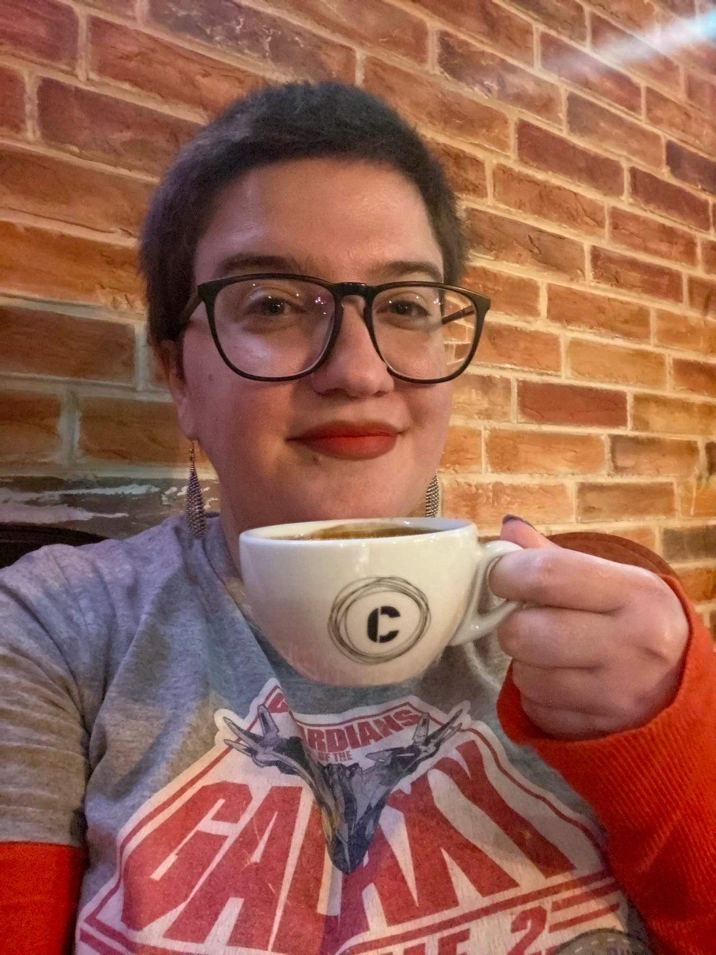 The author sits on a velvet sofa with a cup of coffee in her hand. She is a short-haired brunette with large glasses and a "Guardians of the Galaxy" T-shirt. There is a reddish brick wall behind her. She is looking at the camera and smiling. The author sits on a velvet sofa with a cup of coffee in her hand. She is a short-haired brunette with large glasses and a "Guardians of the Galaxy" T-shirt. There is a reddish brick wall behind her. She is looking at the camera and smiling.