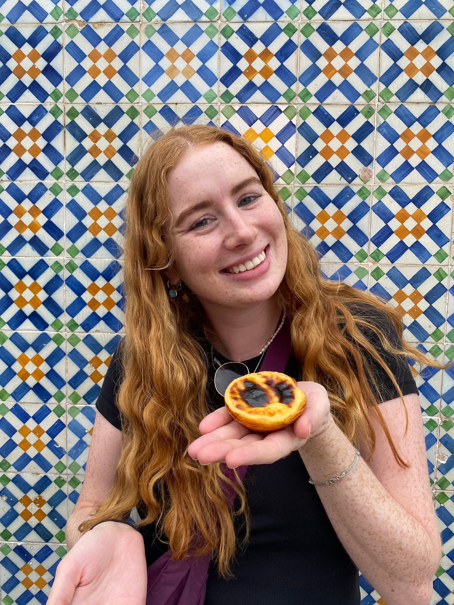 A girl with long red hair holds a pastel de nata in front of a wall covered in blue, yellow, green and white portuguese tiles A girl with long red hair holds a pastel de nata in front of a wall covered in blue, yellow, green and white portuguese tiles
