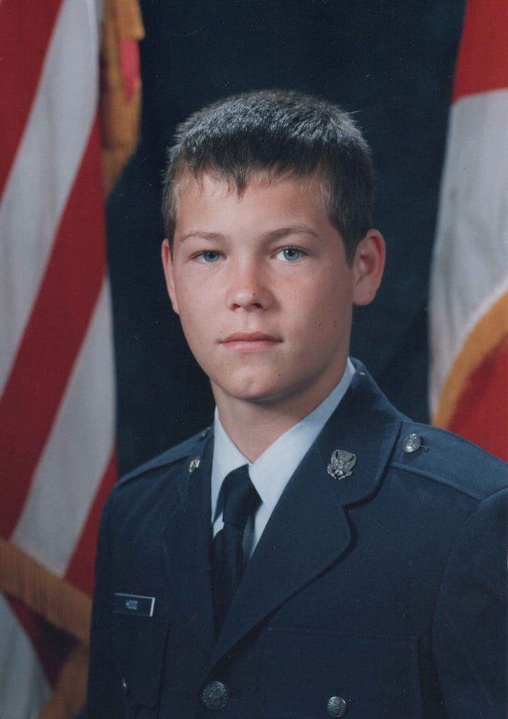 Portrait of a young Edward Hood in a dark military dress uniform with U.S. flags in the background and Blue Star Memorial Highway sign honoring the U.S. Armed Forces beneath a large moss-draped tree by a roadside.