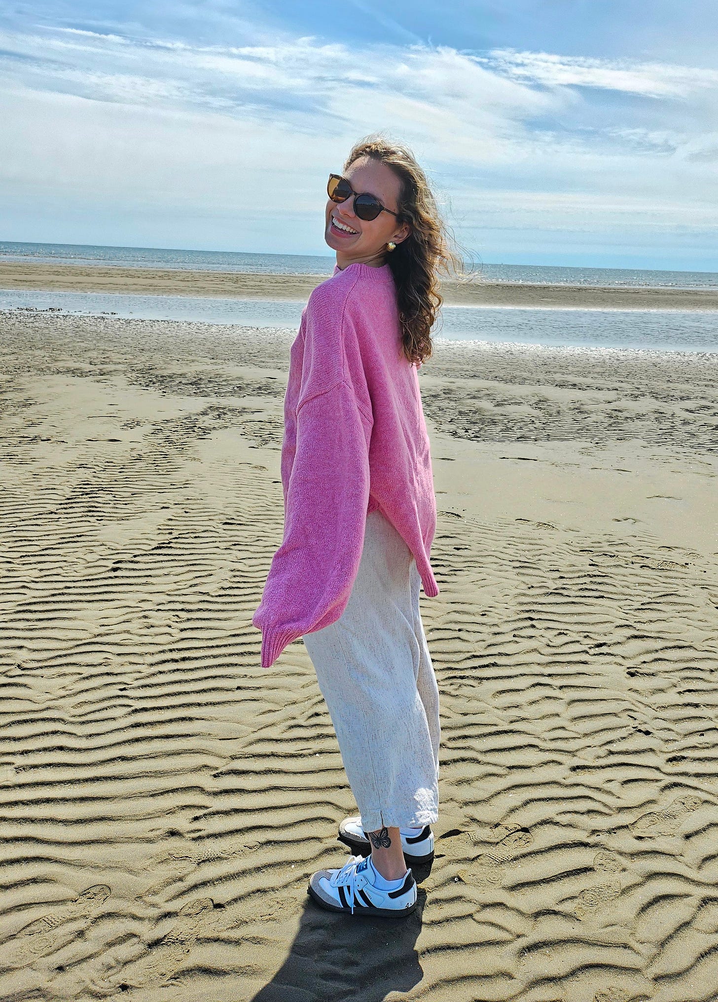 A woman wearing a bright pink sweater, white linen pants, and sunglasses smiles as she looks over her shoulder while walking on a sandy beach. The sky is wide and blue with soft clouds, and gentle ripples in the sand stretch out toward the ocean in the background.