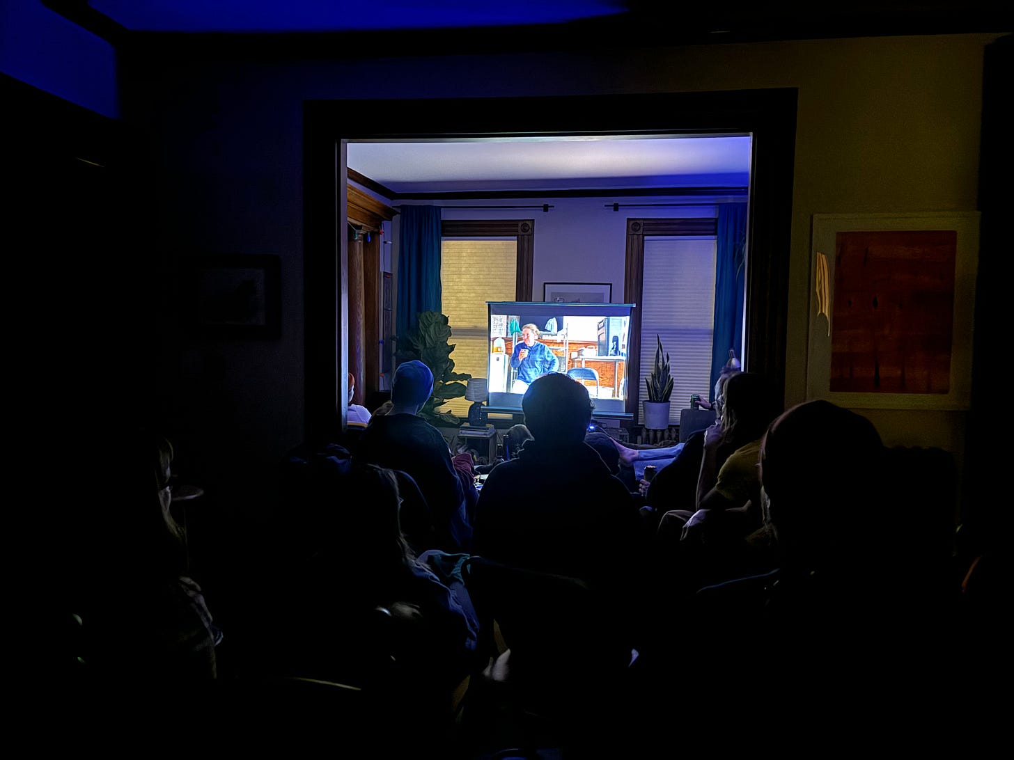 photo of a dark room with a tv on in the front illuminating people sitting on chairs throughout the space.