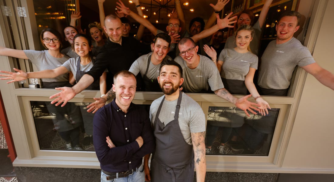 A group of people stood outside and inside a restaurant with their arms outspread smiling A group of people stood outside and inside a restaurant with their arms outspread smiling