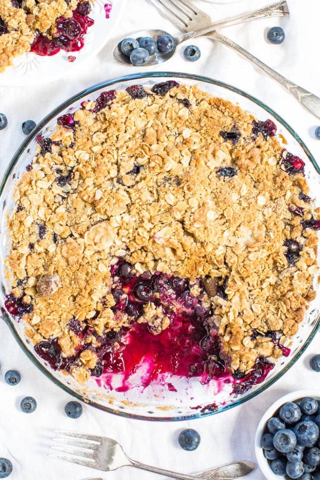 A freshly baked blueberry crumble in a serving dish alongside scattered blueberries and forks on a white background. A freshly baked blueberry crumble in a serving dish alongside scattered blueberries and forks on a white background.
