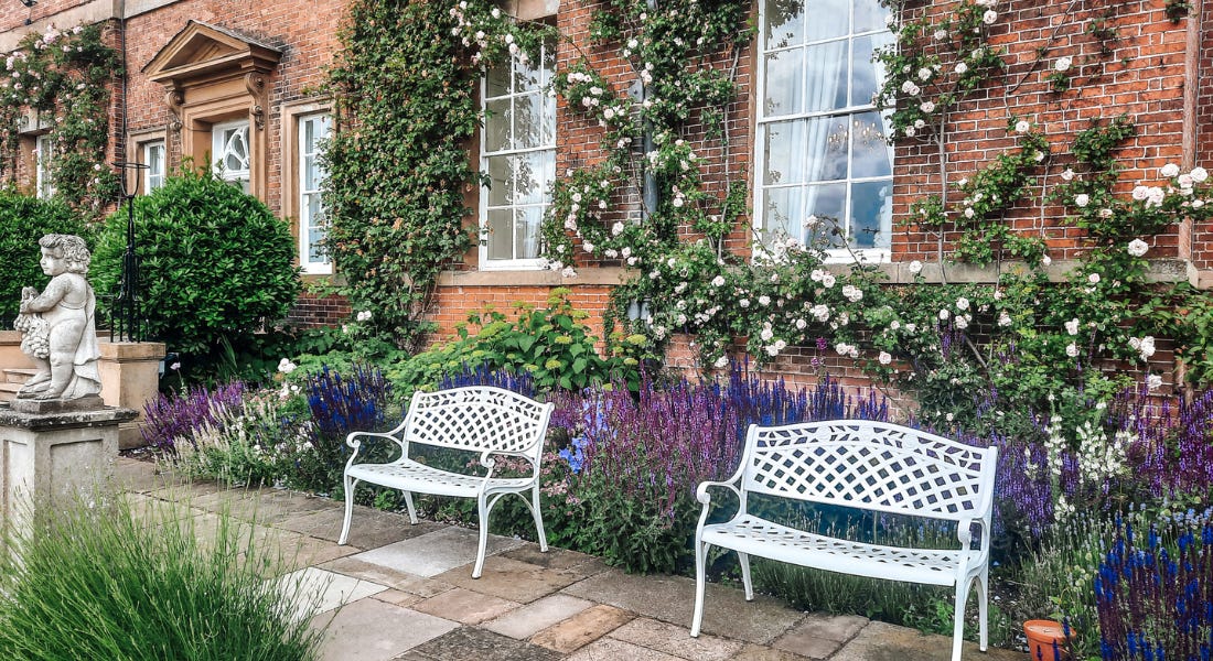 A garden with two white iron benches, purple flowers, and a brick building behind it A garden with two white iron benches, purple flowers, and a brick building behind it