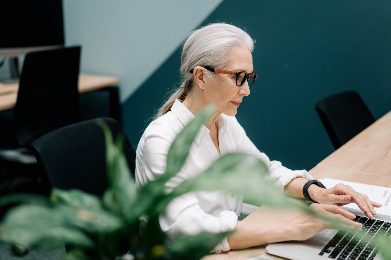 An older woman sits with her hands on a laptop keyboard in a group working space with a turquise and teal striped wall behind her.