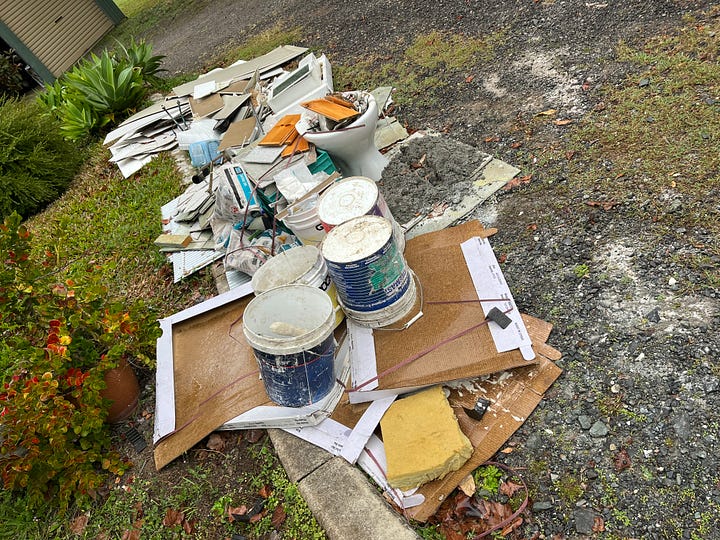 four photos, the first one shows a dated bathroom with dark floor tiles and a shower with a big monstera plant in front of it. The next photo shows the same space with new whitish grey floor tiles just laid. The third photo is an outdoor shot with a pile of garbage that includes: an old toilet, ripped up tiles and other debris from a housing demolition. The final photo is of a front porch with a yellow screen door in the center. On one side is a tile cutting machine on a drop cloth with many other tools of the trade and on the opposite side is a pile of wood trim and boxes of tiles.
