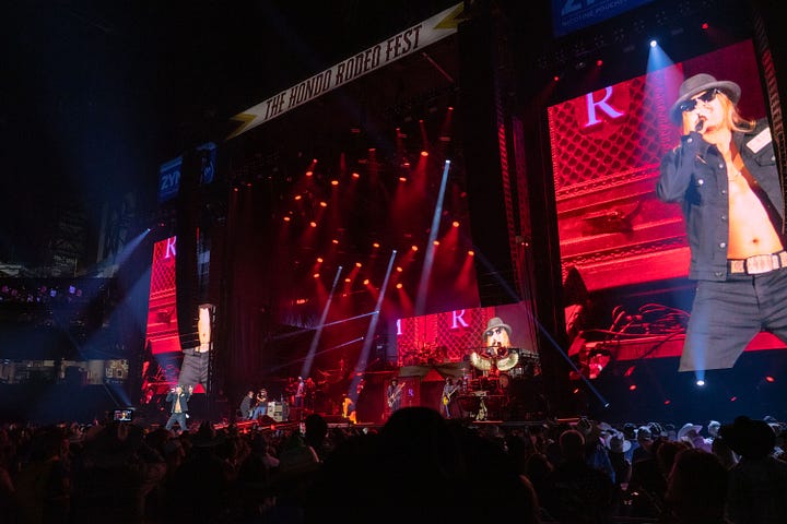 Kid rock is wearing a black open face button down, black hat and black sun glasses singing on stage with his band while the crowd dances to his music