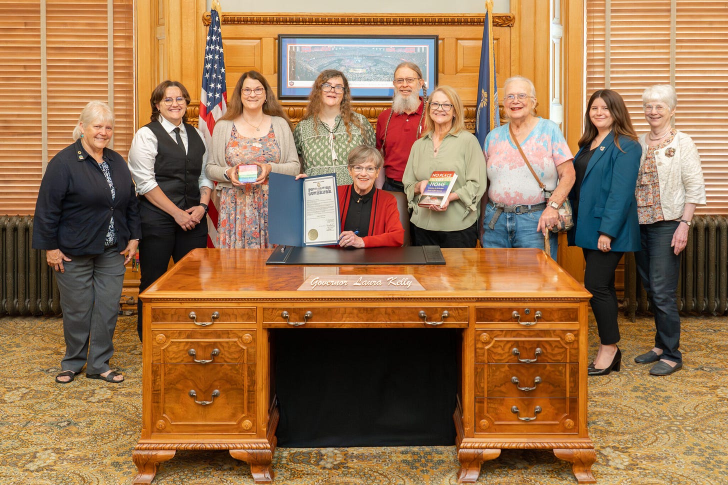 A woman behind a desk holds up a proclamation, while advocates stand behind her. A woman behind a desk holds up a proclamation, while advocates stand behind her.