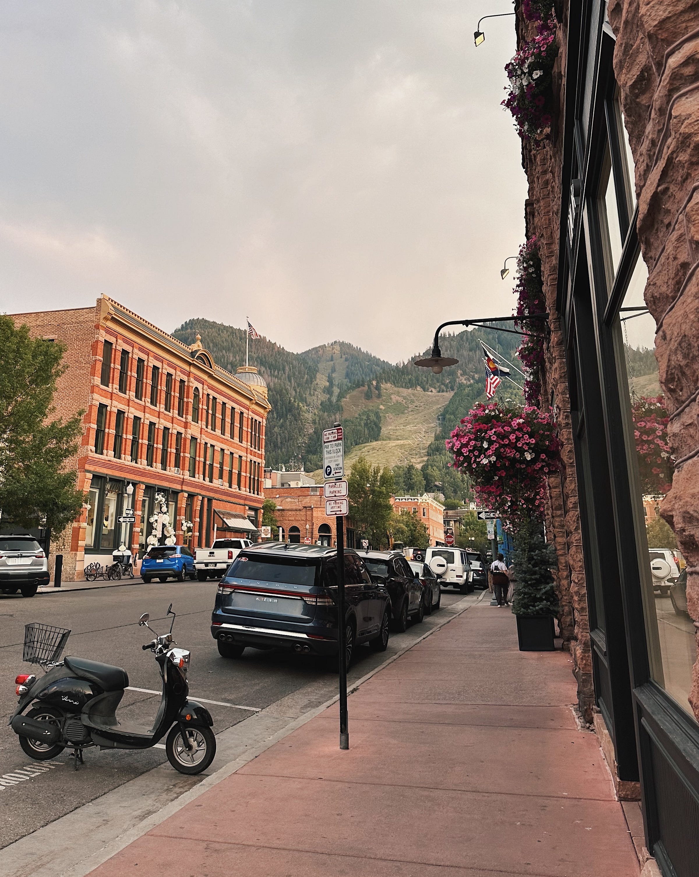 Downtown Aspen street lined with brick storefronts, hanging flowers, and parked cars, with a green mountain rising in the distance.