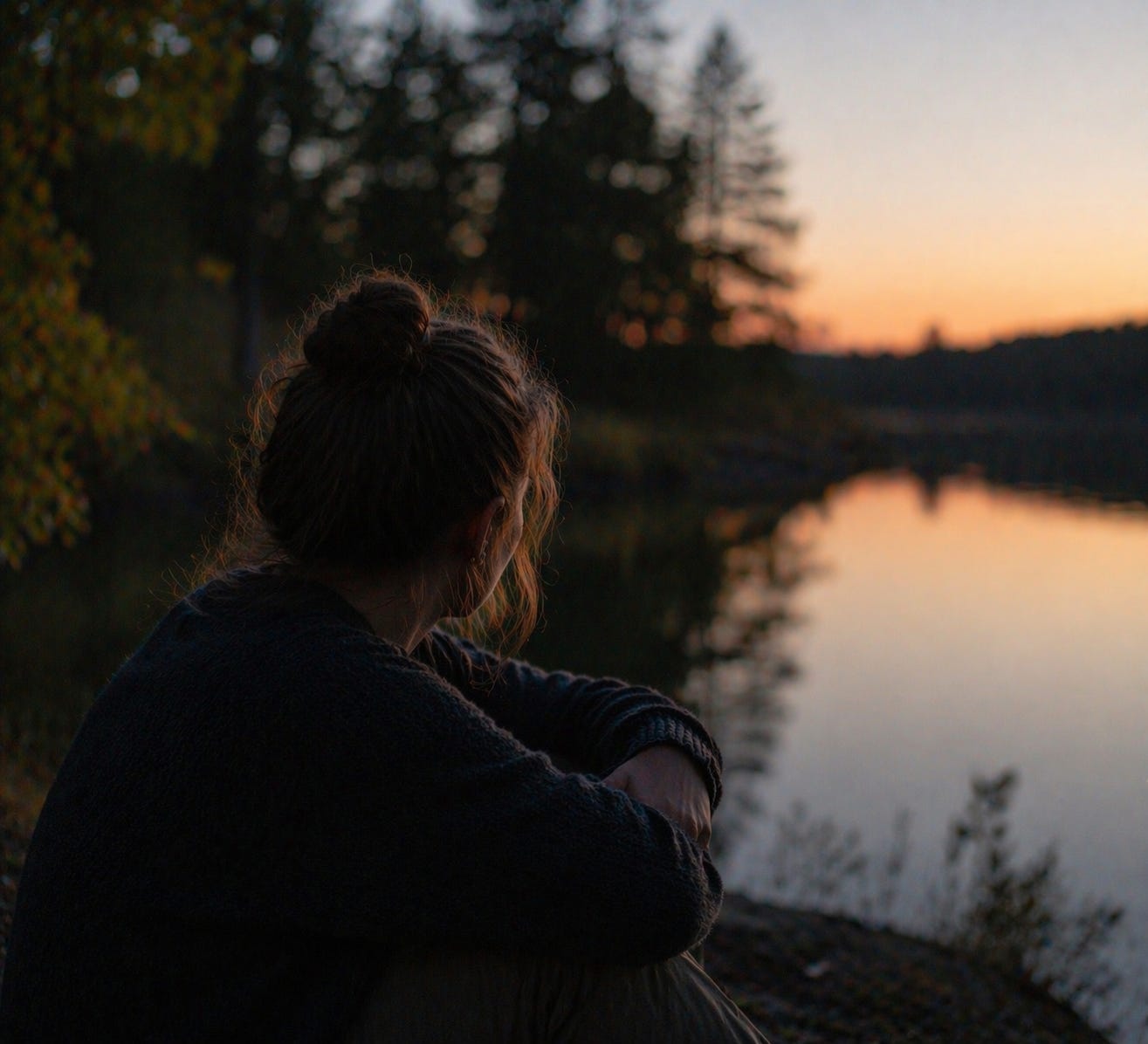 A person sits quietly by a river at dusk, watching the fading light reflect across the water as the surrounding landscape softens into shadow. 