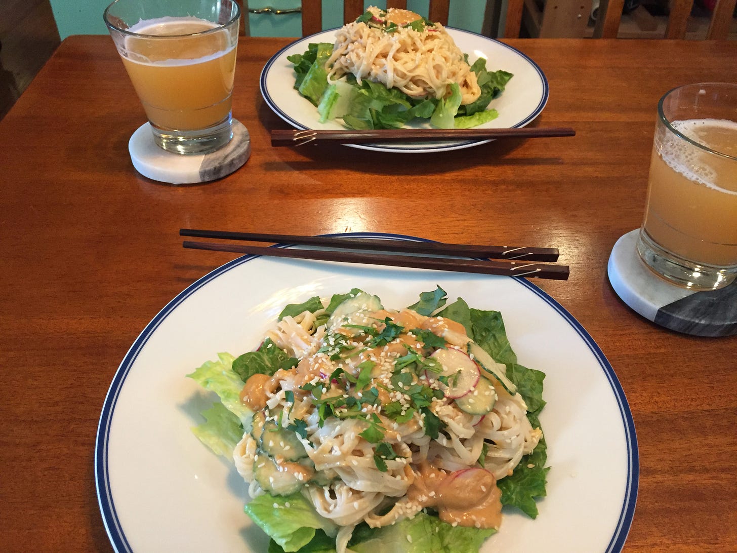 Two plates sit with chopsticks resting on their edges, and in the centre, a pile of rice noodles, cucumber, and radish slices in peanut sauce on a bed of lettuce. Two glass of beer sit on coasters at the sides of the plates.