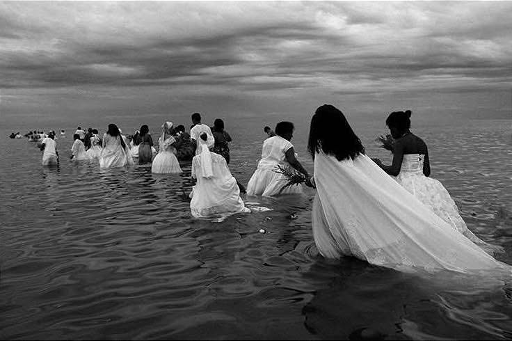 This may contain: a group of women in white dresses wading into the ocean on a cloudy day