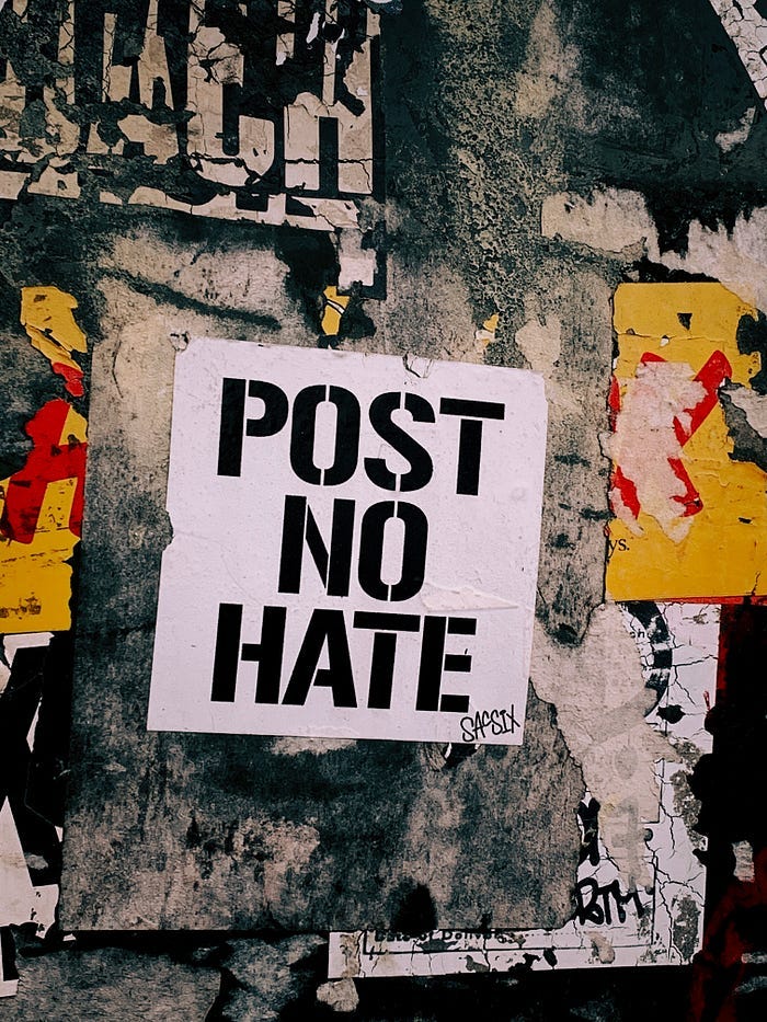 A sign glued to a graffiti’d wall. It reads “Post no hate.”
