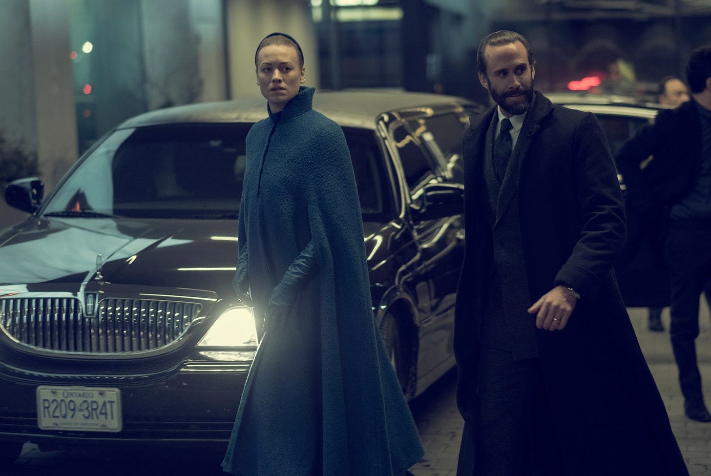 A screenshot from Handmaid's Tale showing Commander Waterford, a tall white man in a suit and dark coat, and Serena Joy, a middle-aged white woman in a blue coat, walking in front of a car with blacked-out windows