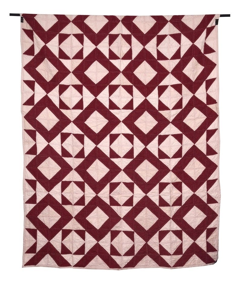 May include: A handmade quilt with a geometric pattern. The quilt features a repeating design of burgundy diamonds and triangles set against a light pink background. The quilt is hanging from a black rod.