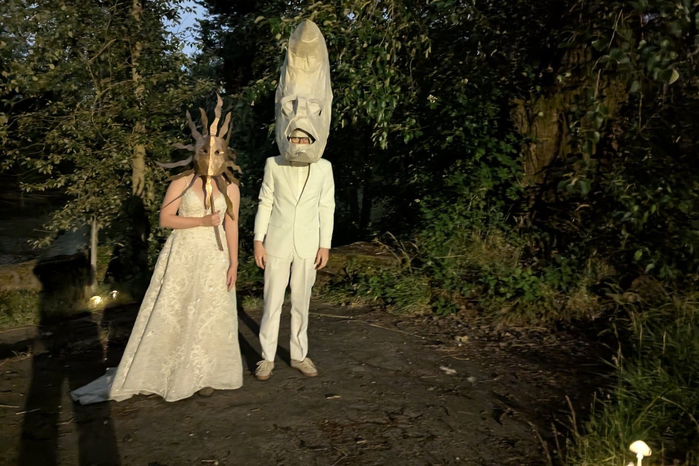 Two people stand in a wooded clearing at dusk wearing elaborate costumes. One wears a white gown with a sunburst-style mask, while the other is dressed in a white suit with a tall papier-mâché moon head. Two people stand in a wooded clearing at dusk wearing elaborate costumes. One wears a white gown with a sunburst-style mask, while the other is dressed in a white suit with a tall papier-mâché moon head.