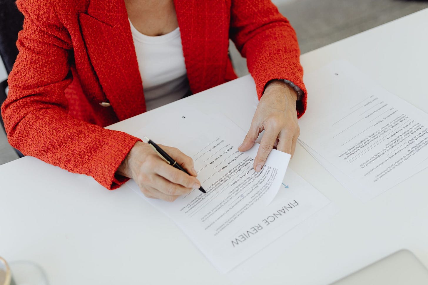 Photo by https://kaboompics.com/ on Pexels Close-up of a businesswoman in a red blazer signing finance documents at an office desk. (stock photo)