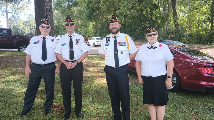 The image shows four adults standing outdoors in front of a maroon Ford Mustang and other vehicles, with large trees and greenery in the background. All four are wearing similar uniforms that include white shirts, black pants or skirt, black shoes, black neckties, and black military-style caps adorned with various insignia and βTALLβ, likely short for Tallahassee. One man is decorated with medals and ribbons. All four individuals are smiling and appear to be part of a formal or ceremonial event, possibly veterans or members of a veterans' organization. No specific names or widely recognizable public figures are evident.