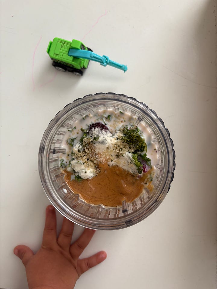 A toddler hand reaching for an unblended smoothie poised beneath a blue and green miniature excavator. A blended smoothie in a ceramic cup next to a stack of books.