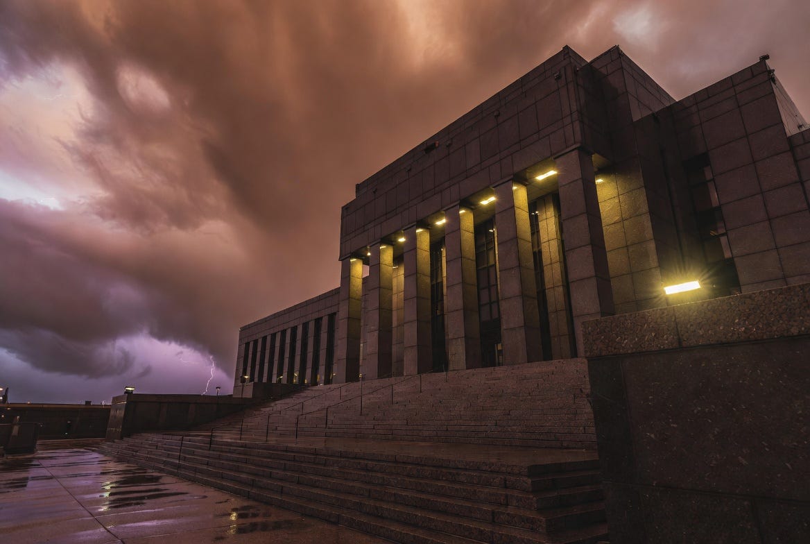 A forbidding neoclassical government building illuminated from within stands under a stormy night sky with lightning striking in the distance, evoking institutional power and impending regulatory confrontation A forbidding neoclassical government building illuminated from within stands under a stormy night sky with lightning striking in the distance, evoking institutional power and impending regulatory confrontation