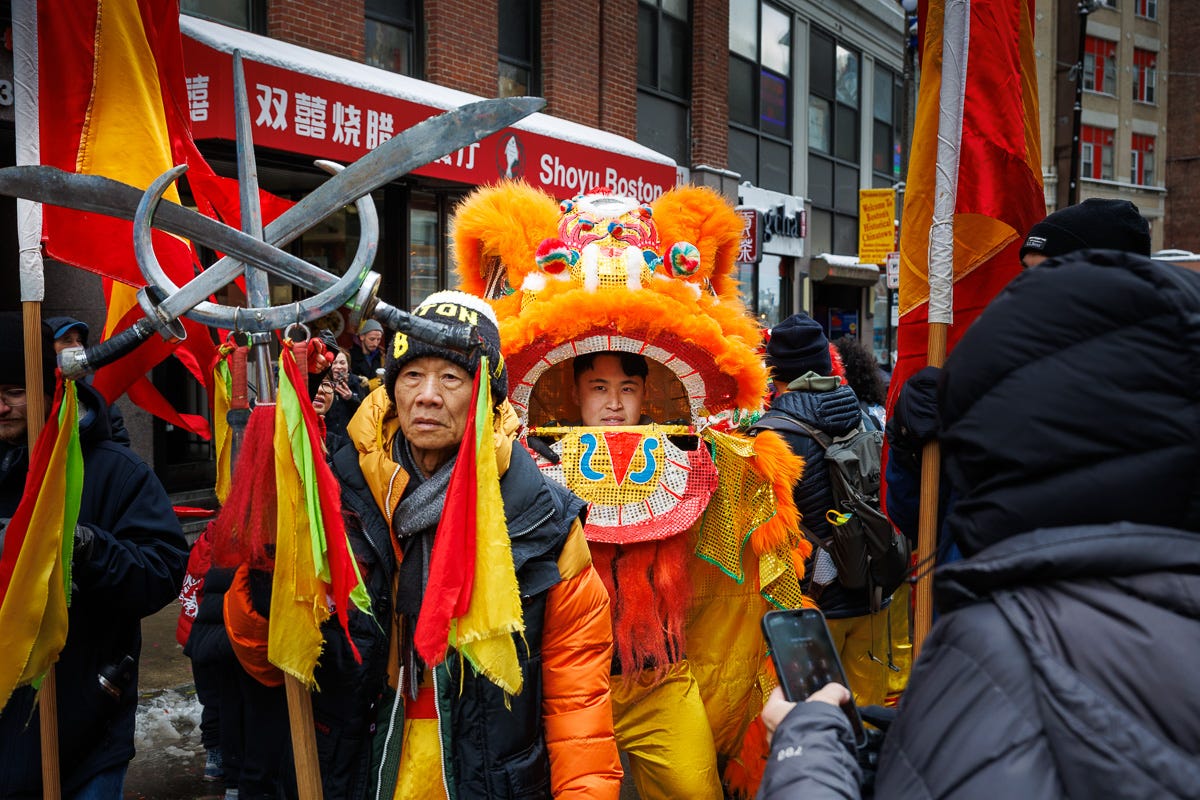 Close-up of a lion dancer and his team during a Chinese New Year parade in Boston’s Chinatown, surrounded by flags and red-orange costume details. Close-up of a lion dancer and his team during a Chinese New Year parade in Boston’s Chinatown, surrounded by flags and red-orange costume details.
