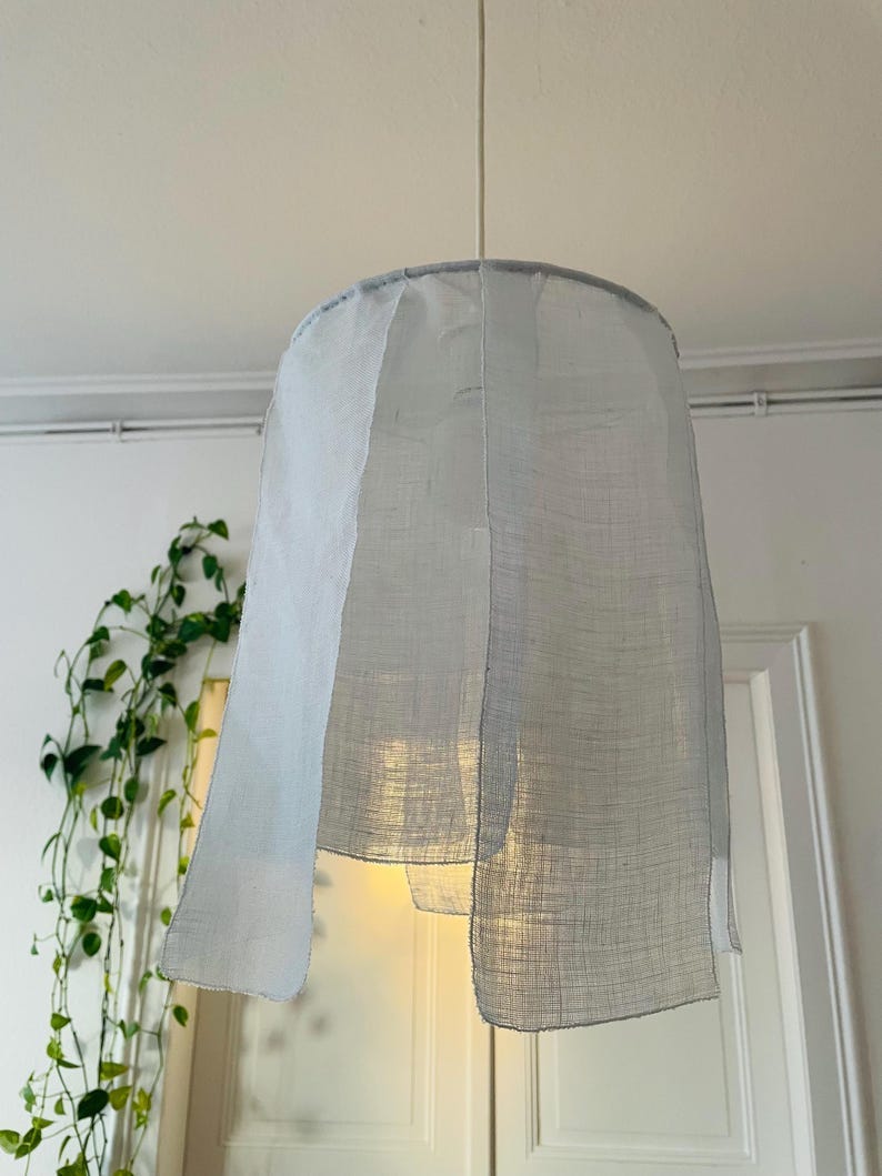 May include: A light blue, fabric lampshade hanging from a white ceiling. The lampshade has a cylindrical shape with a slightly flared bottom. The fabric appears to be semi-transparent, allowing light to filter through. The lampshade is suspended by a thin white cord.