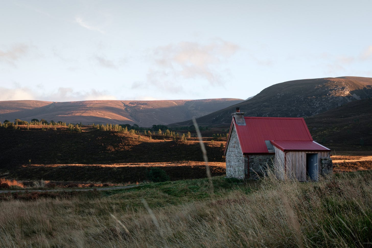 A moody sunset photograph of Ryoan Bothy, a small stone former farmer’s cottage nestled in the Cairngorms. The dark silhouette of the bothy contrasts with the fading sky, framed by vast open hills and golden light on the horizon.