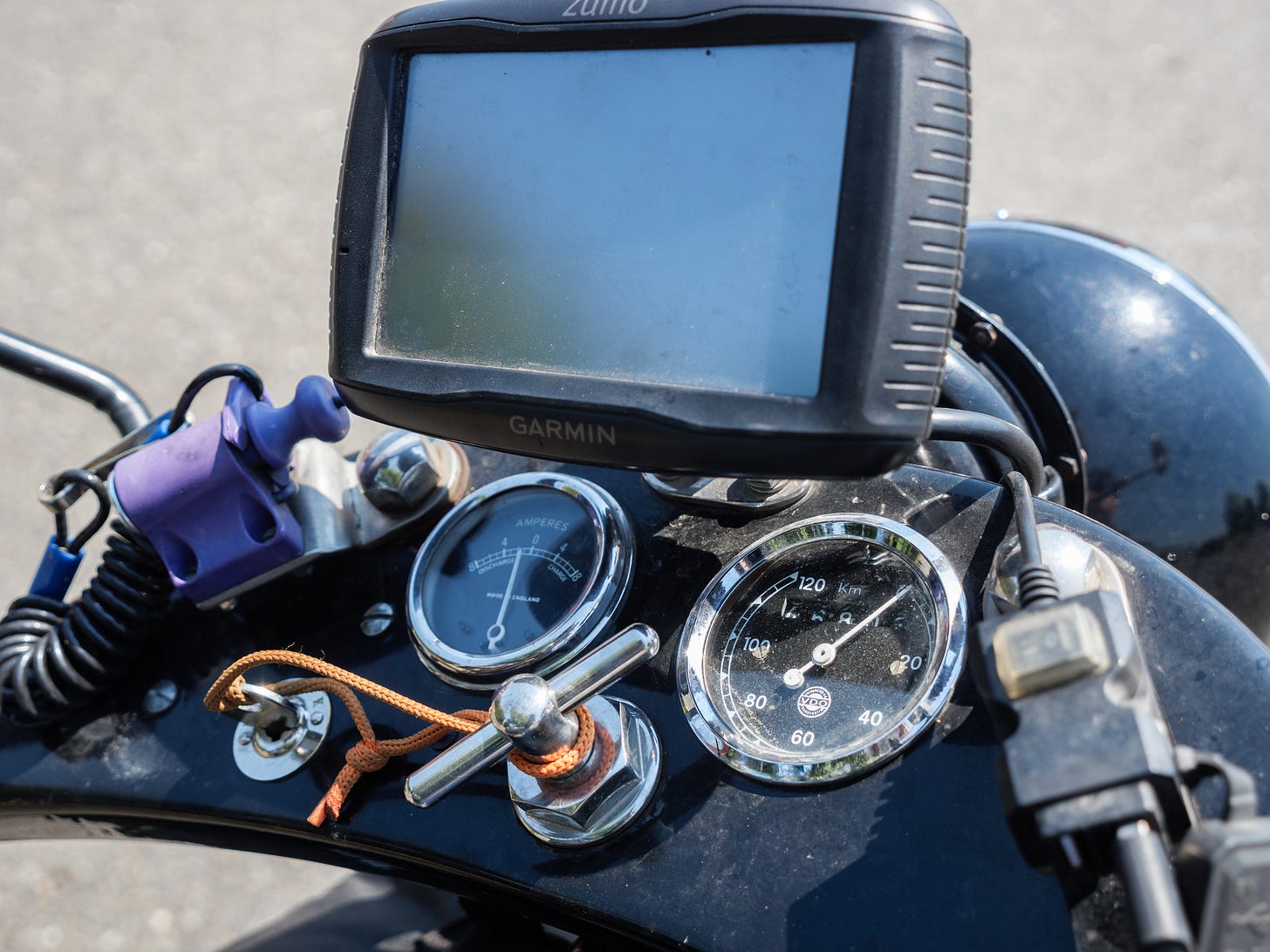 Photo of the dashboard of the old 1930s Nimbus motorcycle, with two, simple gaugues for speed and what is probably engine temperature. The two gauges have black faces, simple white graphics, and chrome trim rings. To the left is a simple ignition with a small key inserted. The key has an orange cord/loop on the end. The loop is secured to the center locknut of the top triple clamp, which features a lever that can be turned. Ahead of the old dials is a more modern Garmin Zumo GPS unit with a large screen, not on. Ahead and below the Garmin is the black headlight nacelle. On the left is a plastic faded purple device of unknown function with a plastic spring attached. On the right is what appears to be a USB connection.