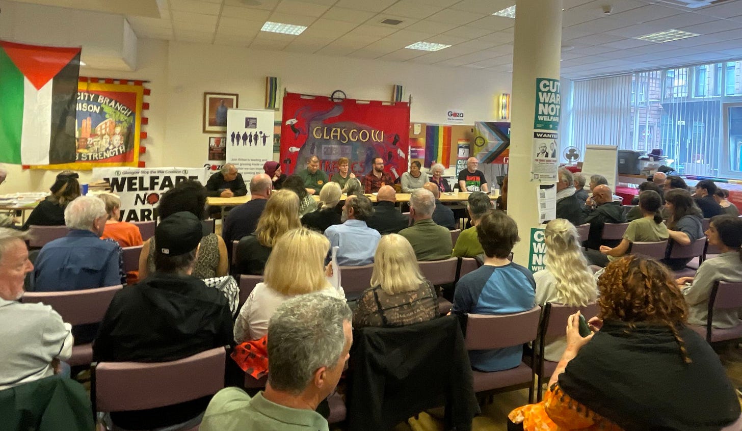 A photo of a jam-packed room of attendees at the Unison Glasgow offices on Bell Street. The panel is sat at the front and there are a number of trade union banners, as well as Palestine and pride flags, adorning the walls.