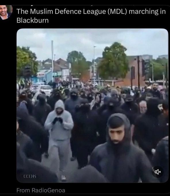The image depicts a large crowd of people, many wearing dark hoodies and face coverings, marching through a street in Blackburn. The setting includes urban elements like trees, buildings, and traffic lights, with the group appearing organized and dense. The post text from Kirsty C references the "Muslim Defence League (MDL)" and suggests the march occurred across major UK cities and towns in summer 2024, resembling an "army." No platform watermarks are visible, and the composition is clear and well-lit, capturing the intensity of the gathering.
