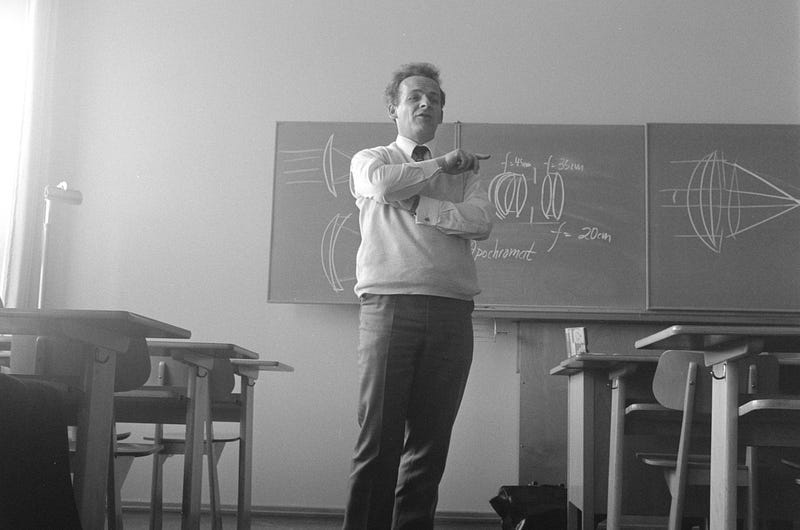 A black and white image of a male teacher in collar, tie and jumper stood in front of a blackboard, pointing as if he might actually know something, which he might not actually do.
