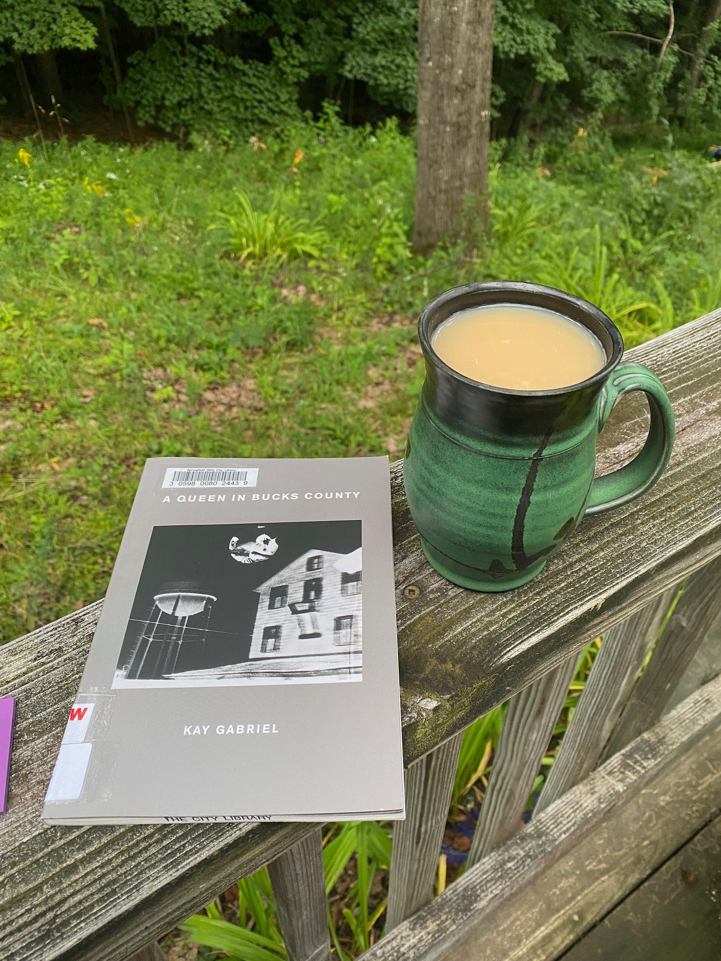 A green ceramic mug of tea and A Queen in Bucks County by Kay Gabriel sit on my porch railing.